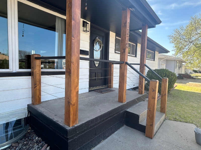 Custom front porch in a modern, industrial style. Stained wooden posts, black handrail, and metal cable wires running the length of the remodeled porch.