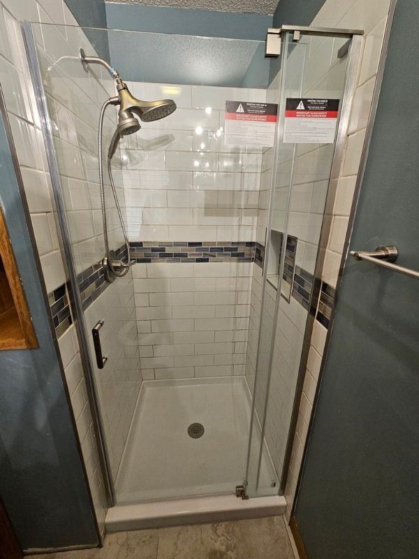 Shower remodel project featuring large, white tile, blue and grey accent tile, a silver shower head, and swinging glass door.